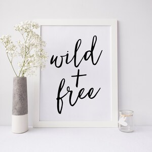 K&ouml;nnte beinhalten: Wei&szlig;er gerahmter Druck mit schwarzer Kalligraphie, die "wild + free" auf wei&szlig;em Hintergrund lautet. Der Druck liegt auf einer wei&szlig;en Oberfl&auml;che mit einer Vase mit wei&szlig;en Blumen und einem durchsichtigen Glasgef&auml;&szlig;.