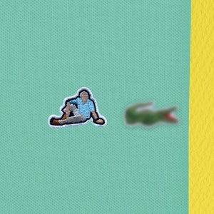 Può includere: Una polo blu chiaro con un logo di coccodrilo verde ricamato sul lato destro del petto. Il coccodrilo è rivolto a destra e ha la bocca rossa.