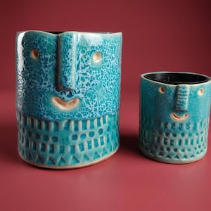 Op de afbeelding: Twee cilindervormige keramische vaten in een levendige turquoise glazuur met een gebarsten textuur. Elk stuk heeft een gestileerd gezicht met een verhoogde neus, kleine ogen en een gebogen mond, met een geometrisch patroon rond de basis.