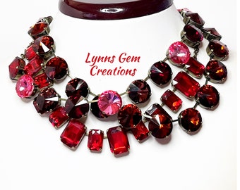 May include: A multi-layered necklace with large red and pink crystal stones. The necklace is made with antique brass findings and features a variety of shapes and sizes of crystals. The necklace is called "Lynns Gem Creations".