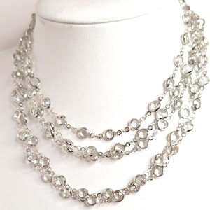 May include: A multi-strand necklace with clear, round crystals set in a silver-toned metal. The necklace is displayed on a white mannequin bust. The crystals are evenly spaced along the strands, creating a sparkling effect.