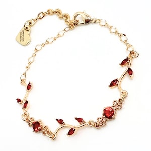 Puede incluir: Pulsera dorada con diseño de vid y detalles de piedras rojas. La pulsera incluye un dije en forma de corazón con el texto "Lynna Gem Creations". Cierre de mosquetón.