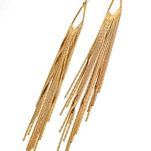 Puede incluir: Un par de pendientes colgantes dorados. Cada pendiente presenta una parte superior geométrica con un diseño largo y con flecos. La borla está hecha de muchos hilos finos y texturizados que caen en cascada. Los pendientes están sobre un fondo blanco.
