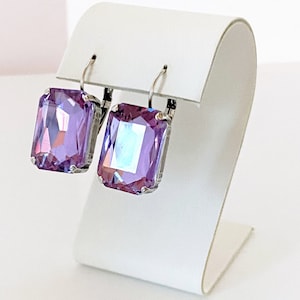 Può includere: Un paio di orecchini d'argento con una pietra preziosa quadrata di colore viola chiaro. Gli orecchini sono esposti su un supporto bianco.