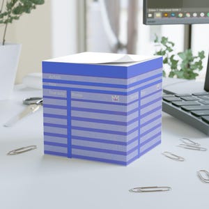 May include: A square blue sticky note cube featuring a printed library card design with horizontal lines and headers for Artist, Title, Due date, and Name. A small crown icon is printed on the side, and the top sheet is slightly curled at the corner.