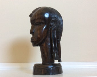 May include: A dark brown wooden sculpture of a person's head. The sculpture is carved in a realistic style and features detailed facial features. The sculpture is mounted on a small wooden base.