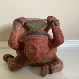 May include: A terracotta sculpture of a person holding a pot above their head. The sculpture is painted in shades of red, brown, and green.