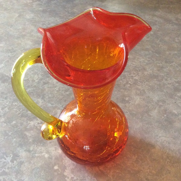 An orange colored hand blown crackle glass pitcher vase with a yellow handle and a fluted sprout, Blenko.