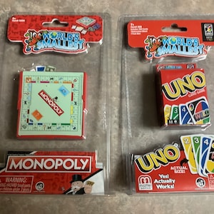 May include: Two packages of the World's Smallest board games. The left package contains a miniature Monopoly board game. The right package contains a miniature Uno card game. Both packages have a red and white background with the game's logo and a warning label.