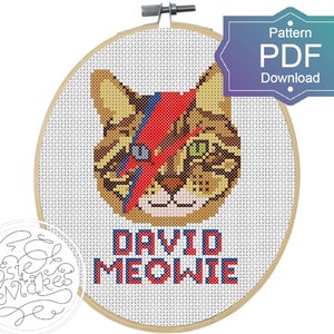 May include: Cross stitch pattern of a cat with a red and blue lightning bolt across its face, with the text "David Meowie" below. The pattern is in a wooden embroidery hoop.