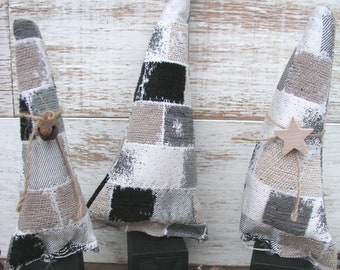 May include: Three decorative fabric Christmas trees in neutral tones of gray, black, and beige, each atop a black block. One tree has a bell, another a wooden star. Two pine cones are in front.