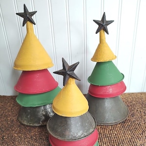 May include: Three decorative Christmas trees made from stacked metal funnels. The trees are painted in yellow, red, green, and gray, and topped with a black star. The trees are arranged on a brown woven surface.