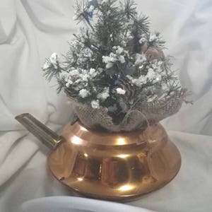 May include: A copper teapot with a rustic burlap base and a small, snowy artificial Christmas tree inside. The tree is decorated with pine cones and blue birds.
