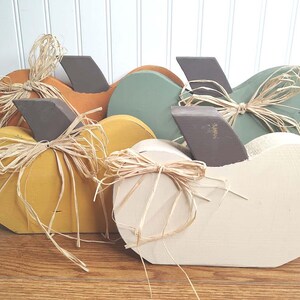 May include: Four wooden pumpkins in various colors, including yellow, orange, green, and white. Each pumpkin has a brown stem and is decorated with a straw bow.