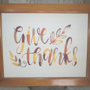 May include: A framed canvas print with the words "give thanks" in a colorful, autumnal font. The words are surrounded by stylized leaves in shades of orange, yellow, and brown.