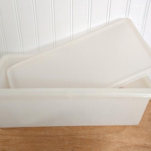 May include: Off-white rectangular plastic container with a matching lid. The lid is partially covering the container. The container is sitting on a wooden surface. The background is a white wall with vertical paneling.