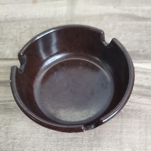 May include: A dark brown ashtray with a rounded shape and three notches for resting cigarettes. The ashtray has a smooth, glossy finish and is made of a solid material. The ashtray is a vintage item.