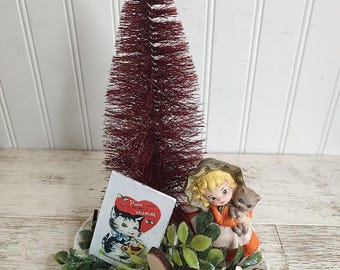 May include: A Valentine's Day themed decorative piece featuring a burgundy bottle brush tree topped with a wooden heart. A small figurine of a girl holding a kitten sits on a plate with greenery, a Valentine's card, and wooden hearts.