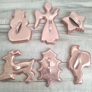 May include: A collection of six rose-gold colored cookie cutters in festive shapes. The shapes include a snowman, an angel, a star, a reindeer, a holly leaf, and Santa Claus. Each cutter has a metal bar for easy use. Perfect for holiday baking.