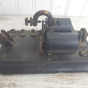 May include: Antique black telegraph machine with brass accents. The device features a lever, multiple terminals, and two cylindrical components. The base is rectangular, and the overall design suggests a vintage communication tool.