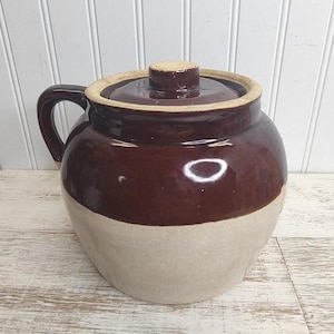 May include: A two-tone, brown and beige ceramic crock with a lid and handle. The crock has a rounded shape and a glossy finish. The lid has a small knob for easy removal. This vintage-style crock is suitable for kitchen storage or decoration.