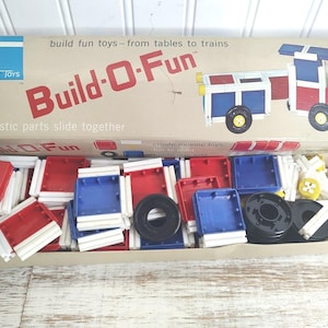 May include: Vintage Tupperware Toys Build-O-Fun set. The box features a red, white, and blue train illustration. Inside are red, blue, and white plastic building blocks, black wheels, and other parts. The box reads "Build fun toys - from tables to trains."