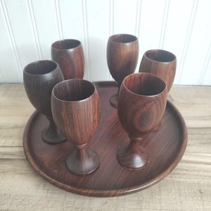 May include: A set of six wooden wine glasses and a matching serving tray. The glasses and tray are made of a dark brown wood with visible grain patterns. The glasses are arranged on the tray, ready for use.