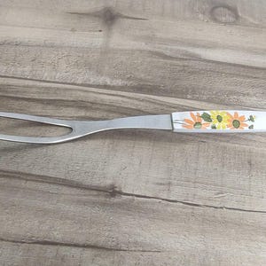May include: A stainless steel carving fork with a white handle decorated with orange and yellow floral designs. The two-pronged fork is designed for serving meats and other dishes. The handle has a hole for hanging.