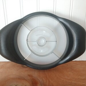 May include: Black apple slicer with a white interior and a clear plastic blade guard. The slicer has a rounded shape with two handles on either side. The slicer is sitting on a wooden surface.