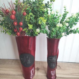 May include: Two red vases crafted from repurposed vinyl records, each featuring a central black record label. The vases are filled with artificial greenery, including stems with red glittered berries on the left and small white flowers on the right.