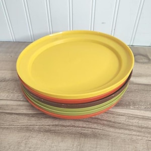 May include: A stack of colorful oval-shaped plates. The top plate is yellow, followed by orange, brown, and green plates. The plates are stacked on a light-colored wooden surface.