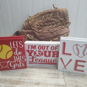 May include: Three wooden signs with red and white lettering and baseball-themed graphics. The signs read "Let's do this girls", "I'm out of your league", and "Love". A brown leather baseball glove is in the background.