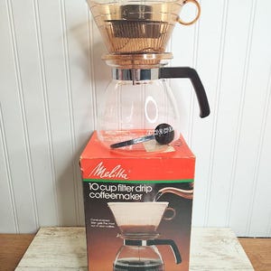May include: A vintage Melitta 10-cup filter drip coffeemaker in a red box. The coffeemaker is made of glass and has a brown plastic filter. The box has a picture of the coffeemaker and the text "Melitta 10 cup filter drip coffeemaker".