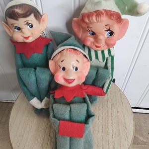 May include: Three elf dolls with painted faces, pointed ears, and felt clothing. Two dolls wear dark green outfits with red collars, while the third wears a light green striped outfit and a matching hat. Each doll has a soft body and a plastic face.