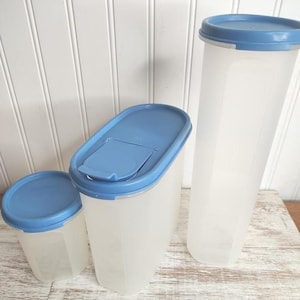 May include: Three clear plastic food storage containers with blue lids. The containers are different sizes and shapes, including a tall, rectangular container and two shorter, oval-shaped containers. The containers are on a light-colored wooden surface.