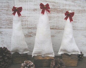 May include: Three white fabric Christmas trees with red bows on top, standing on wooden bases. The trees are arranged in a row on a white wood background.