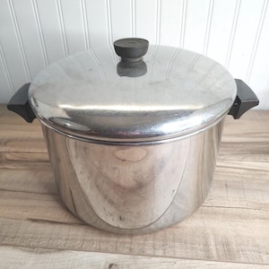 May include: A stainless steel cooking pot with a lid. The pot has a shiny, reflective surface and black handles on either side. The lid has a black knob. The pot is sitting on a wooden surface.