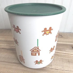 May include: A white cylindrical plastic storage container with a dark green lid. The exterior features a repeating pattern of brown gingerbread people with colorful heart centers and brown gingerbread houses with green chimneys.