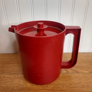 May include: A red plastic pitcher with a lid and a handle. The pitcher is sitting on a wooden surface.