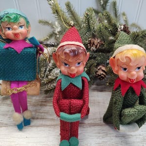 May include: Three vintage Christmas elf ornaments with green, red, and brown felt bodies. The elves have plastic faces with painted features and are wearing green, red, and white felt hats.