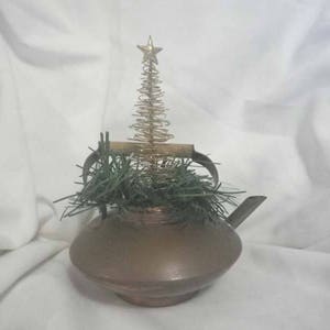 May include: A copper teapot with a gold wire Christmas tree and green pine branches inside. The teapot has a spout and a handle.