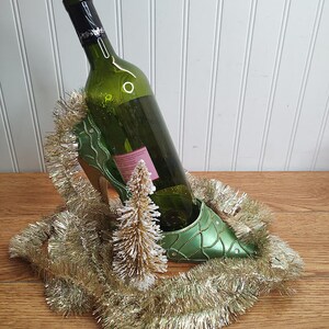 May include: A green glass bottle of wine is in a green high heel shoe shaped wine bottle holder. The holder is surrounded by gold tinsel.