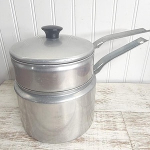 May include: A silver double boiler with a black knob on the lid. The boiler has two separate pots, one nested inside the other. The boiler is made of metal and has a shiny finish.