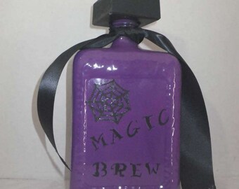 May include: Purple glass bottle with a black lid and a black ribbon. The bottle has a spiderweb design and the words "Magic Brew" written on it.