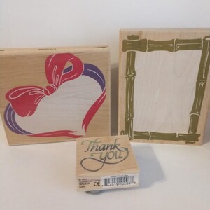 May include: Three wooden rubber stamps. One stamp features a heart with a red bow and purple ribbon. Another stamp features a bamboo frame. The third stamp says "Thank You" in a cursive font.