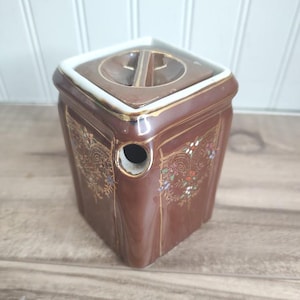 May include: A brown ceramic container with a square shape and a lid. The container has gold trim and floral designs. It has a hole on the side. The lid has a unique design. The container is on a wooden surface.
