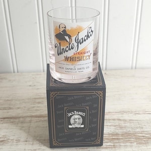 May include: A clear whiskey glass with the text "Uncle Jack's STRAIGHT WHISKEY" and a portrait of a man. The glass sits on a black box with the Jack Daniel's logo and text. The box has gold accents.