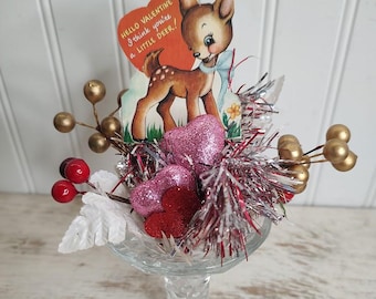 May include: A glass vase filled with red and pink glitter hearts, gold and white sprigs, and a vintage Valentine's Day card featuring a cartoon deer with the text "Hello Valentine I think you're a little deer!"