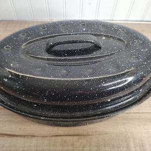 May include: Oval black enamel roasting pan with a lid. The pan has a speckled white pattern and a black handle. The lid has several small holes. The pan is sitting on a wooden surface.