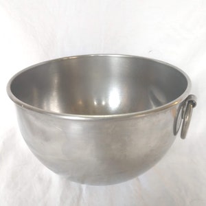 May include: A round, silver-colored stainless steel mixing bowl with a smooth, polished finish. The bowl features a wide, open top rim and a single metal ring handle attached to the side, providing a functional grip for handling the container.